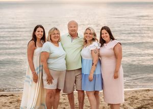 A family of five adults poses together smiling on a sandy beach at golden hour sunset.