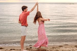 A young couple dances playfully on a sandy beach with calm ocean waves at sunset.