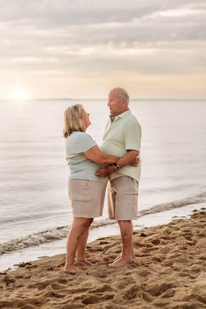 A senior couple holds each other tenderly while standing on a sandy beach at sunset.