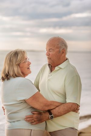 An older couple embraces and gazes into each other eyes on a serene beach at dusk.