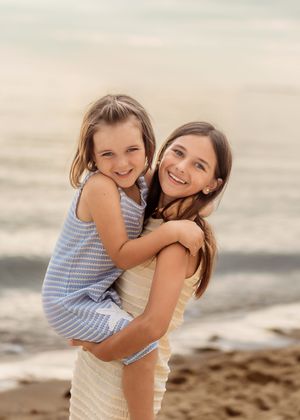 An older girl smiles while giving her younger sister a piggyback ride on a sunny beach.