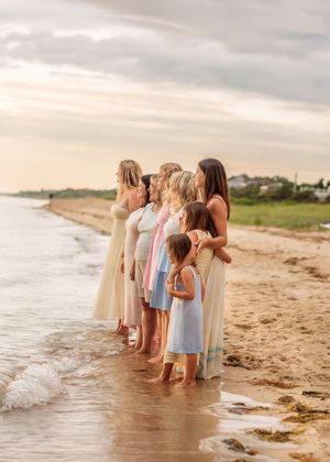 Group of women and girls in pastel dresses standing together at the beach's edge during golden hour.