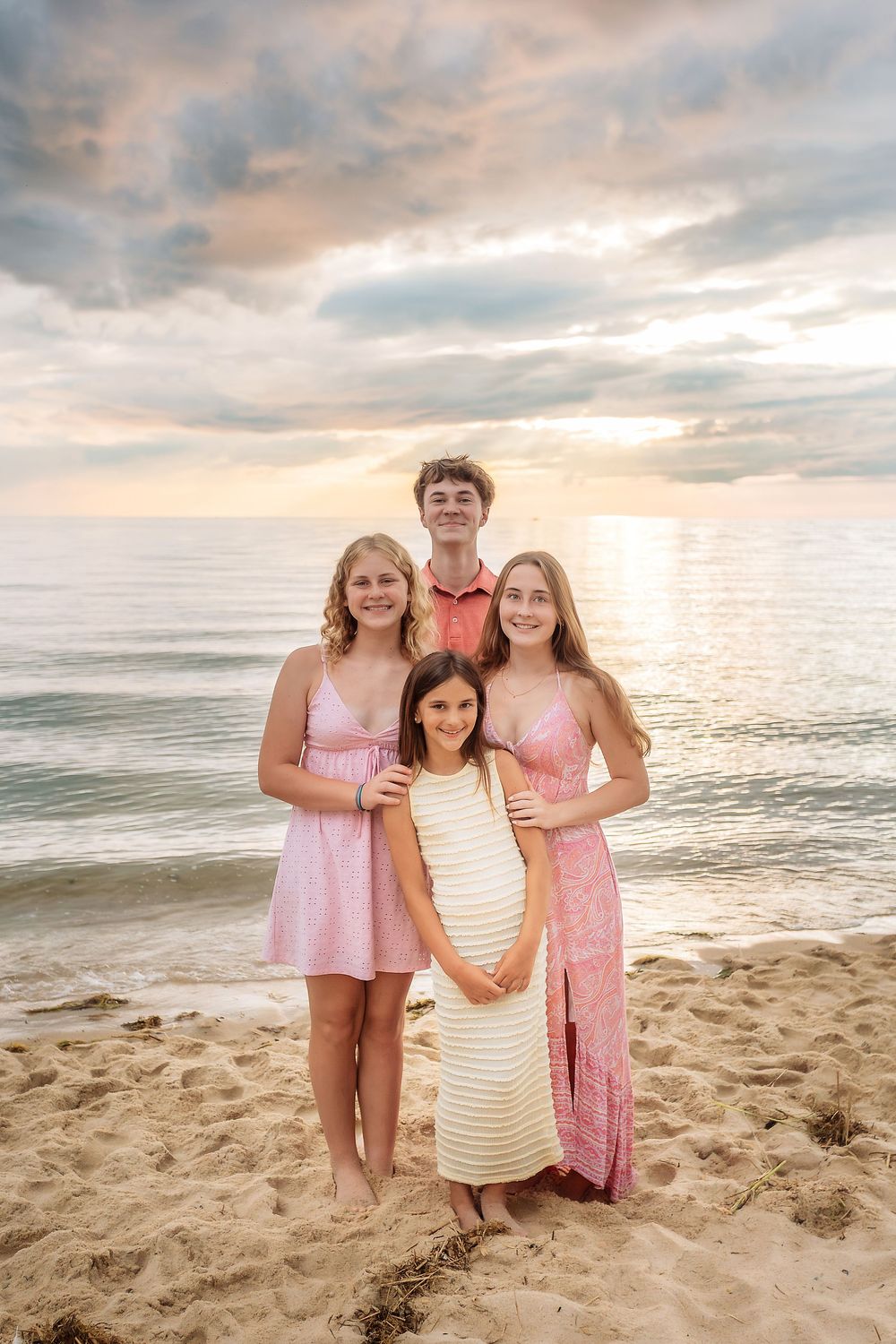 Four siblings pose together on a sandy beach at sunset, wearing pink and white summer outfits.