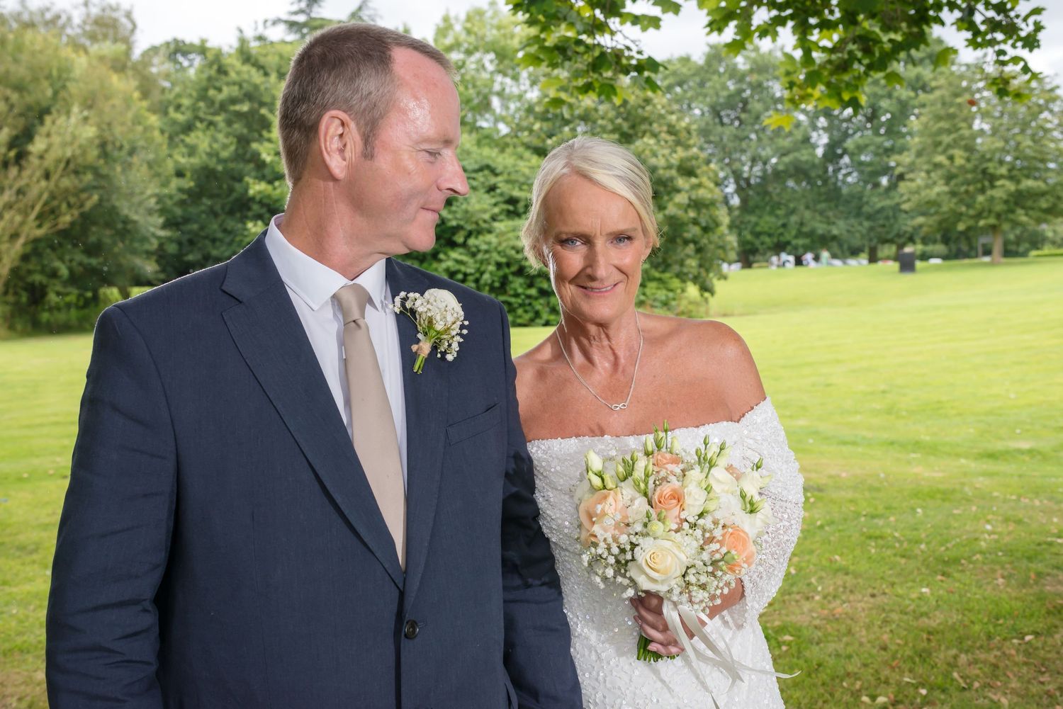 Bride and groom smiling together outdoors at a wedding, bride holding a bouquet of peach and white roses.