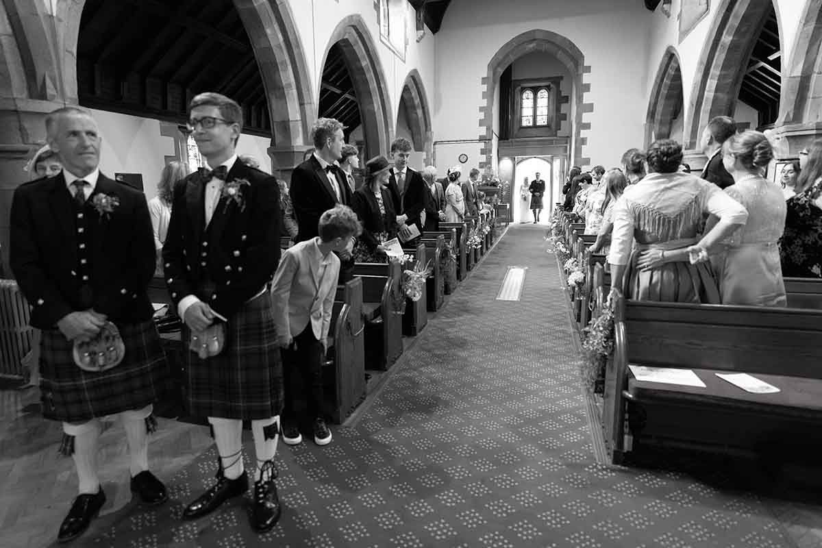 Black and white wedding photo of groomsmen in kilts standing in a church aisle during a ceremony.