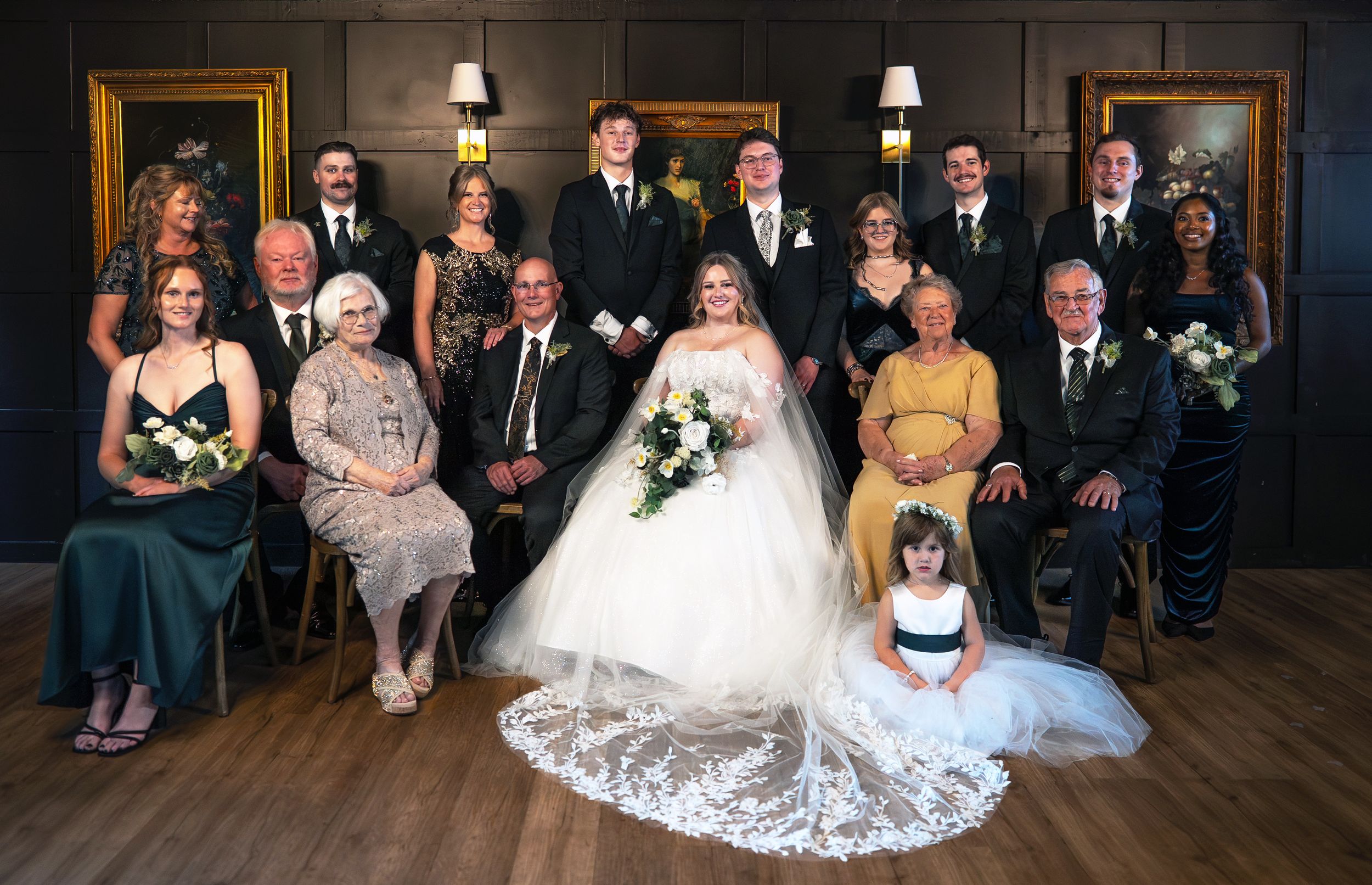 Wedding party poses formally in elegant dark-paneled room, bride in white gown surrounded by family in emerald green attire.