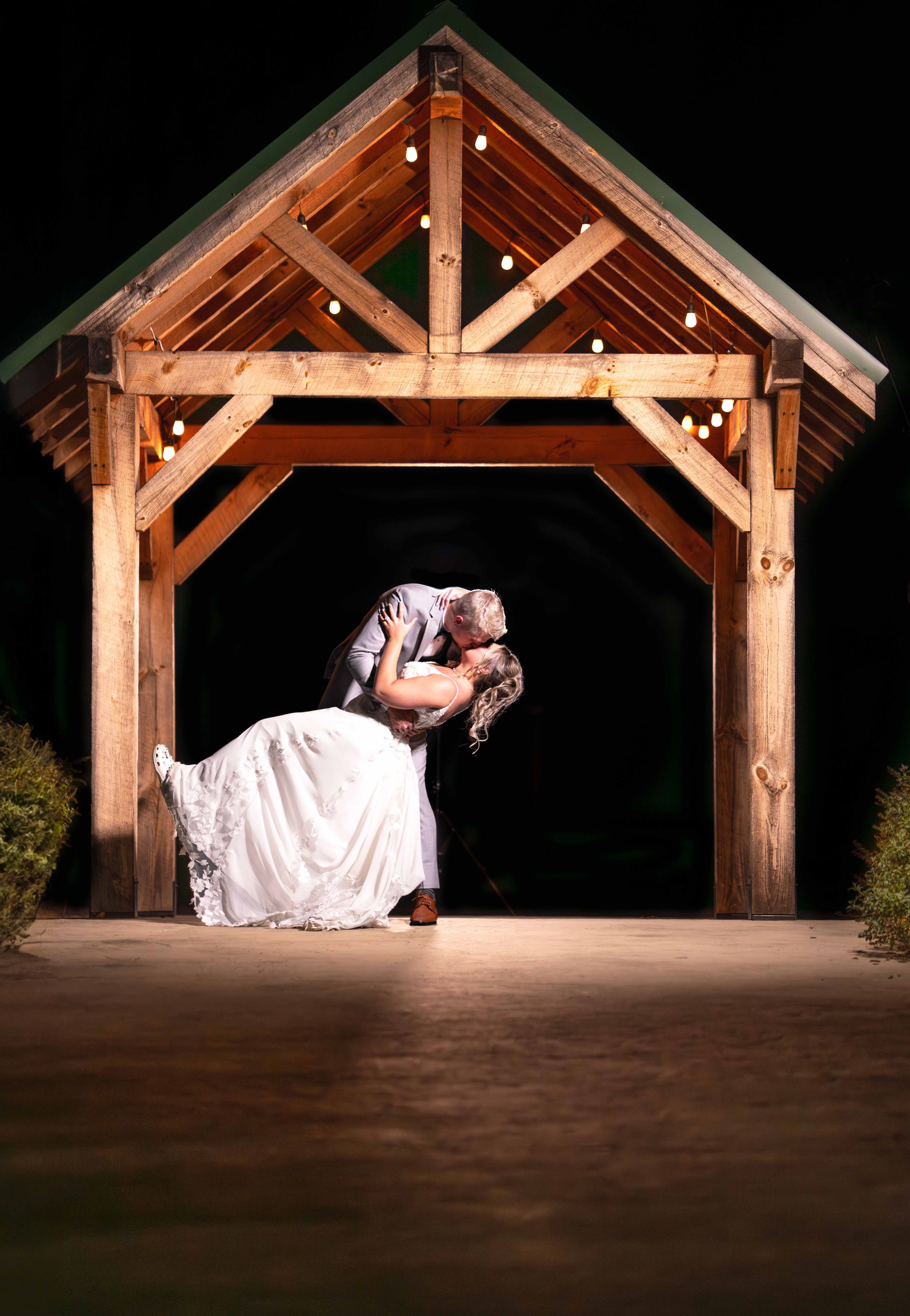 Bride and groom share a romantic dip kiss under a rustic wooden pergola with string lights at night.