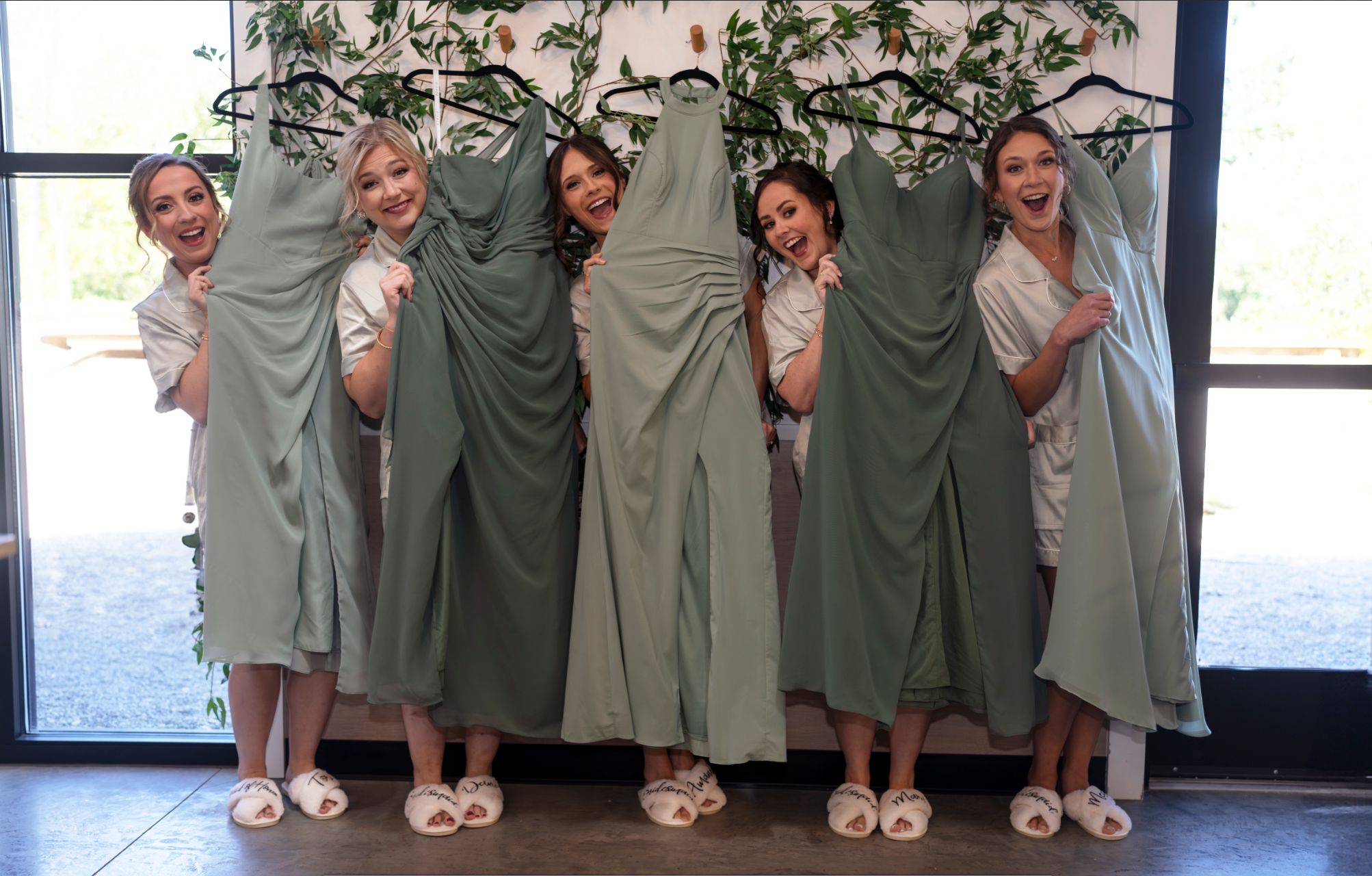Five bridesmaids in robes holding sage green bridesmaid dresses on hangers, smiling together before a wedding.