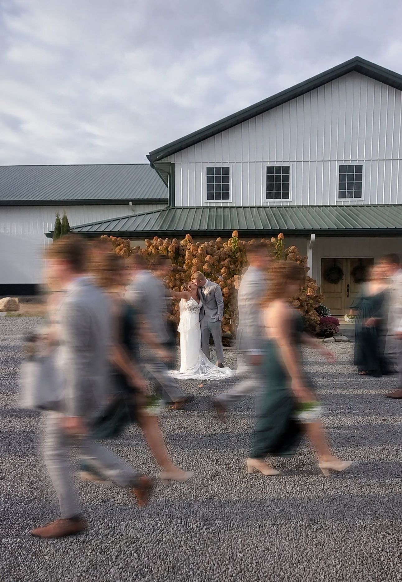Bride and groom pose outside a white barn venue while blurred wedding guests walk past on gravel.