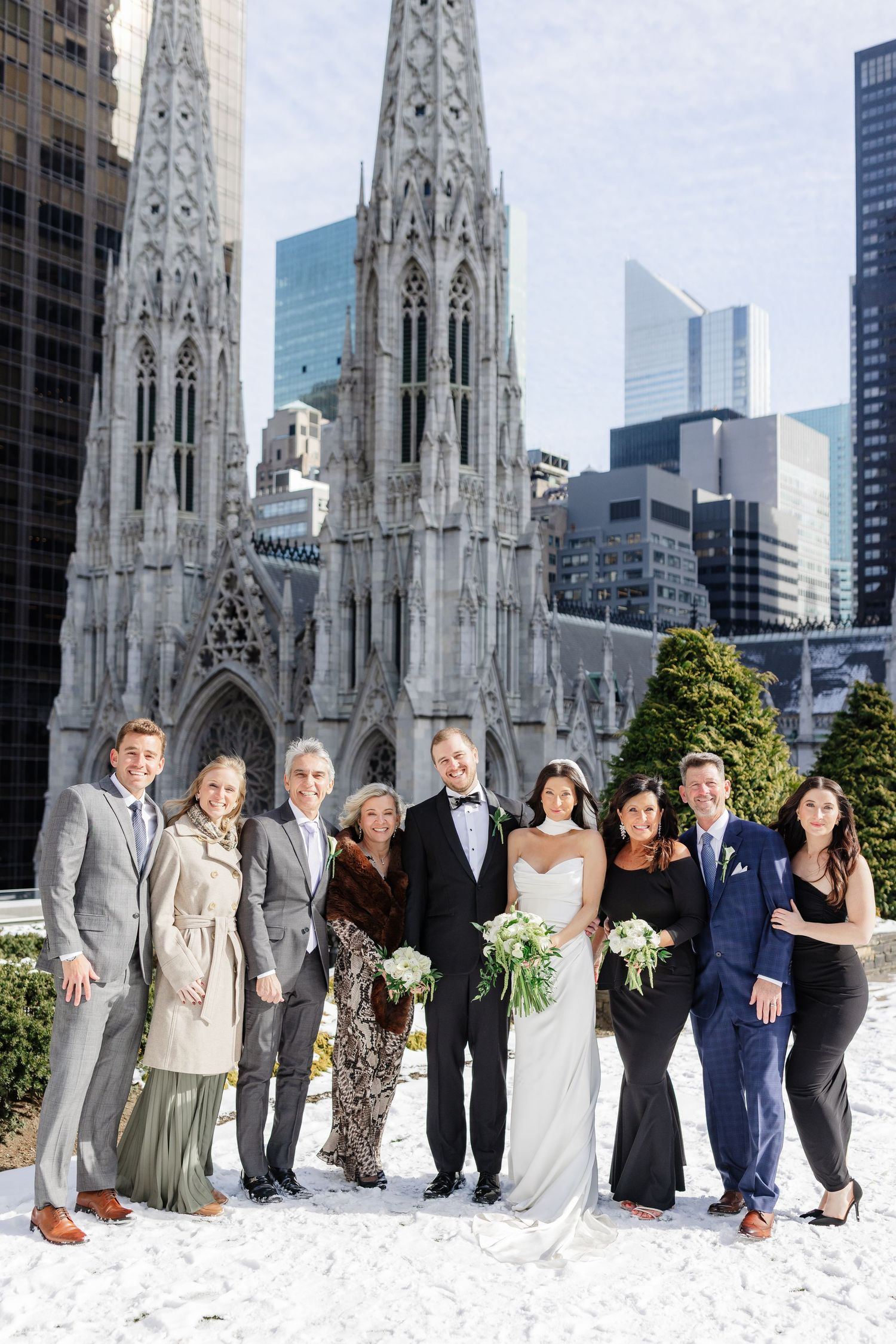 Wedding party poses in snow before St. Patrick's Cathedral NYC spires, bride in white gown with bouquet, groom in tuxedo.
