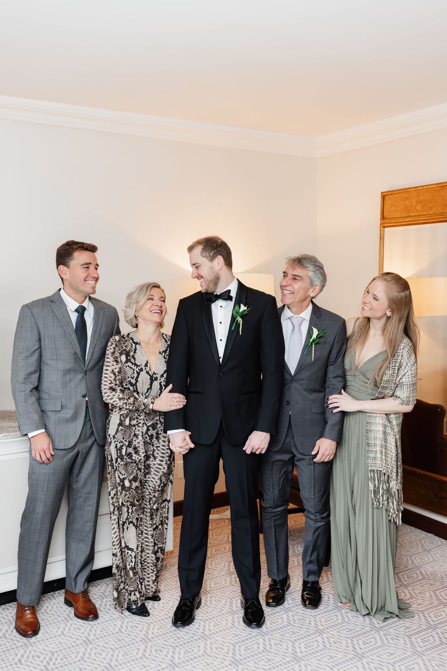 Groom in tuxedo poses with family members in formal attire during wedding day preparations indoors.