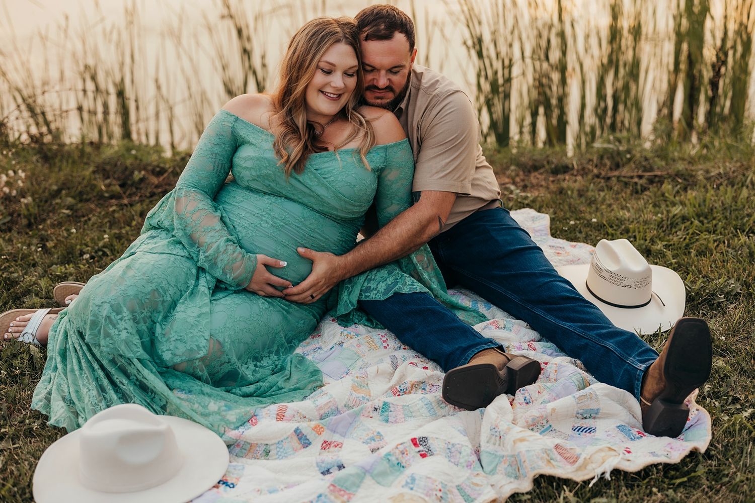 Couple sitting on a quilt by a lake, woman in teal maternity gown cradling baby bump during a maternity photo session.