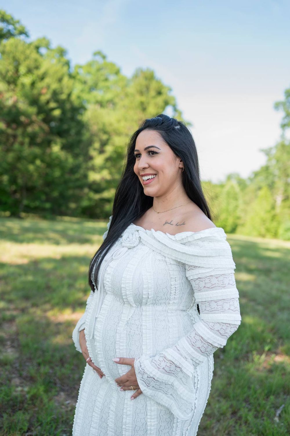 Smiling pregnant woman in white off-shoulder dress holding baby bump outdoors in green field in North Carolina