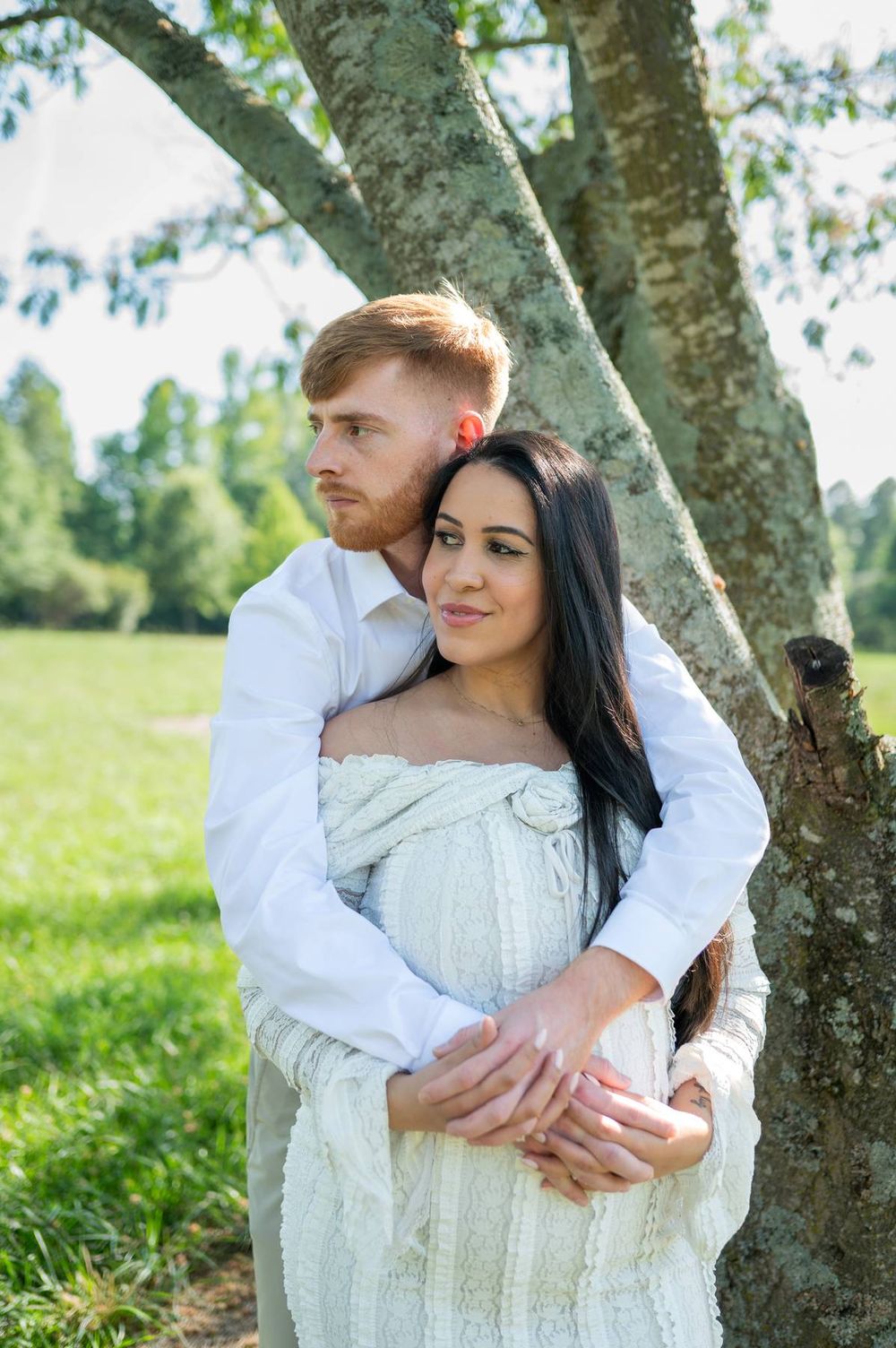 Couple embracing by tree outdoors, man hugging pregnant woman in white knit dress in green park in North Carolina