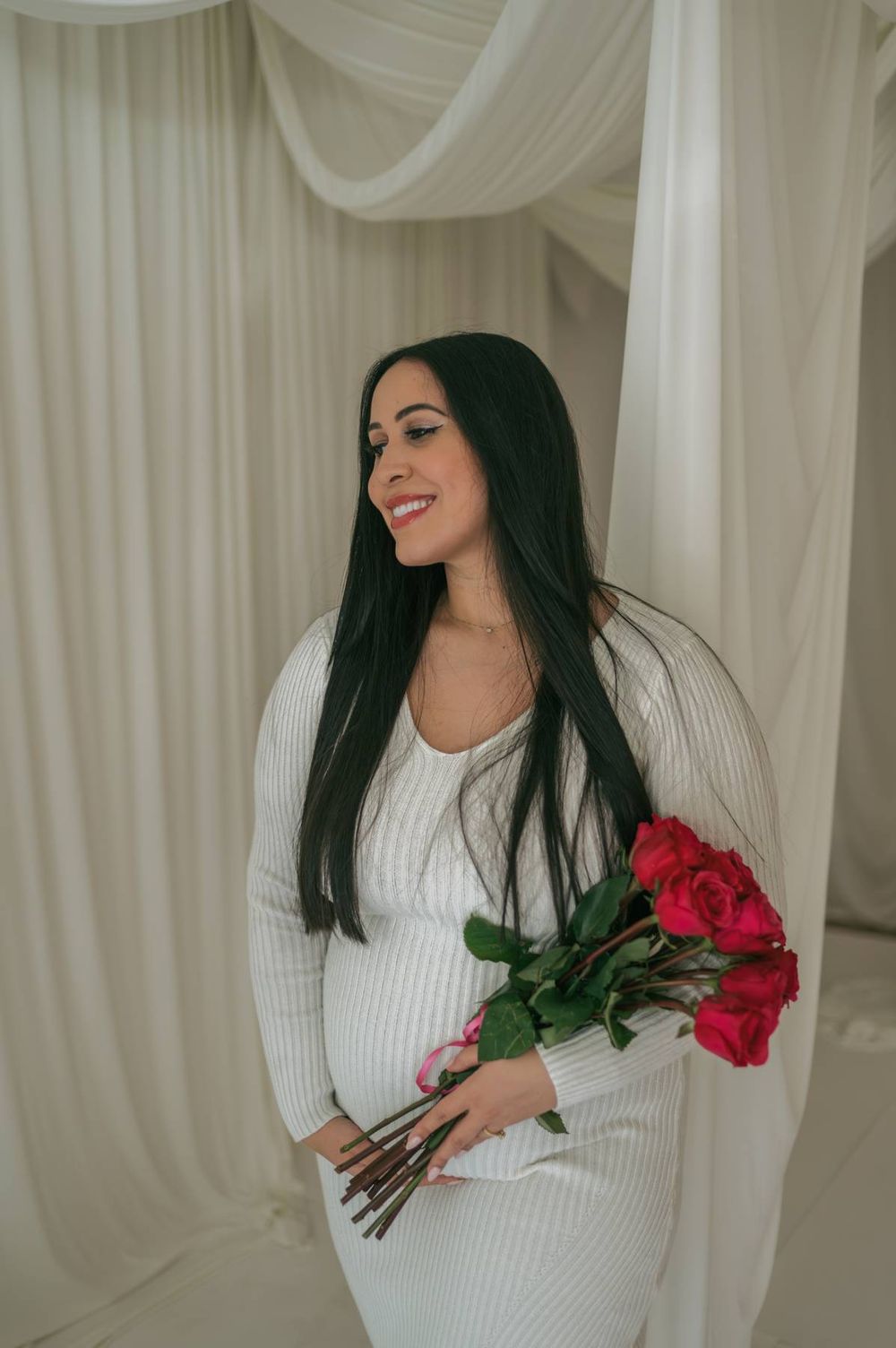 Smiling pregnant woman in white ribbed dress holding bouquet of red roses indoors with white drapes Lexington NC