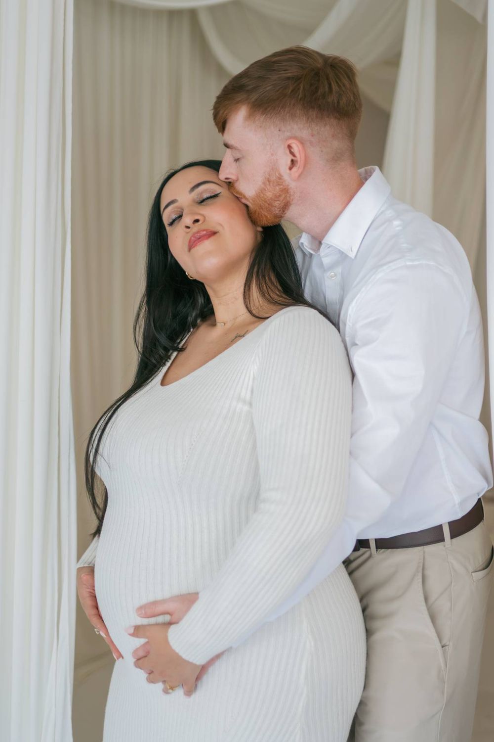 Man kissing pregnant woman's cheek as she smiles with eyes closed, both dressed in white indoors Greensboro NC