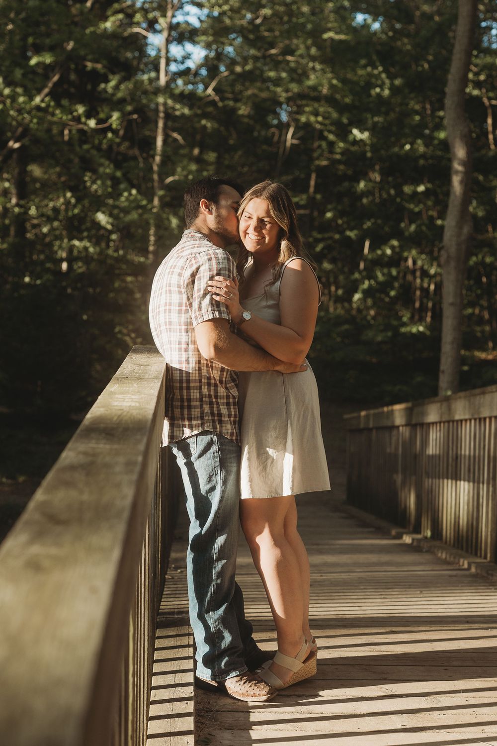 Couple sharing a romantic moment on a wooden bridge surrounded by lush green trees in warm sunlight.
