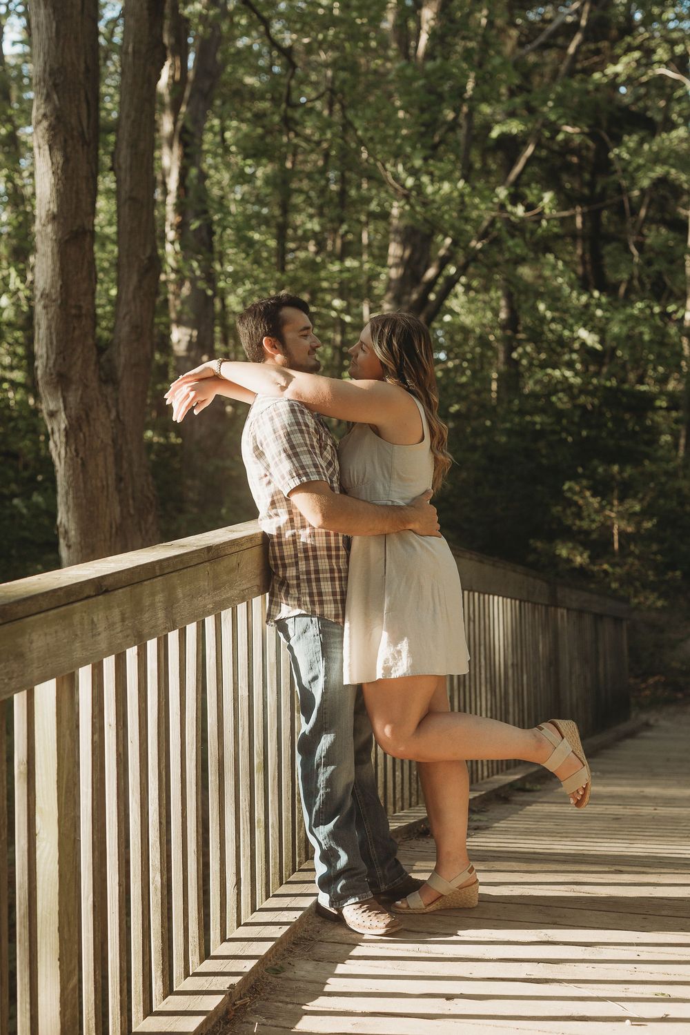 Couple embracing and kissing on a wooden bridge surrounded by lush green trees in a forest setting.