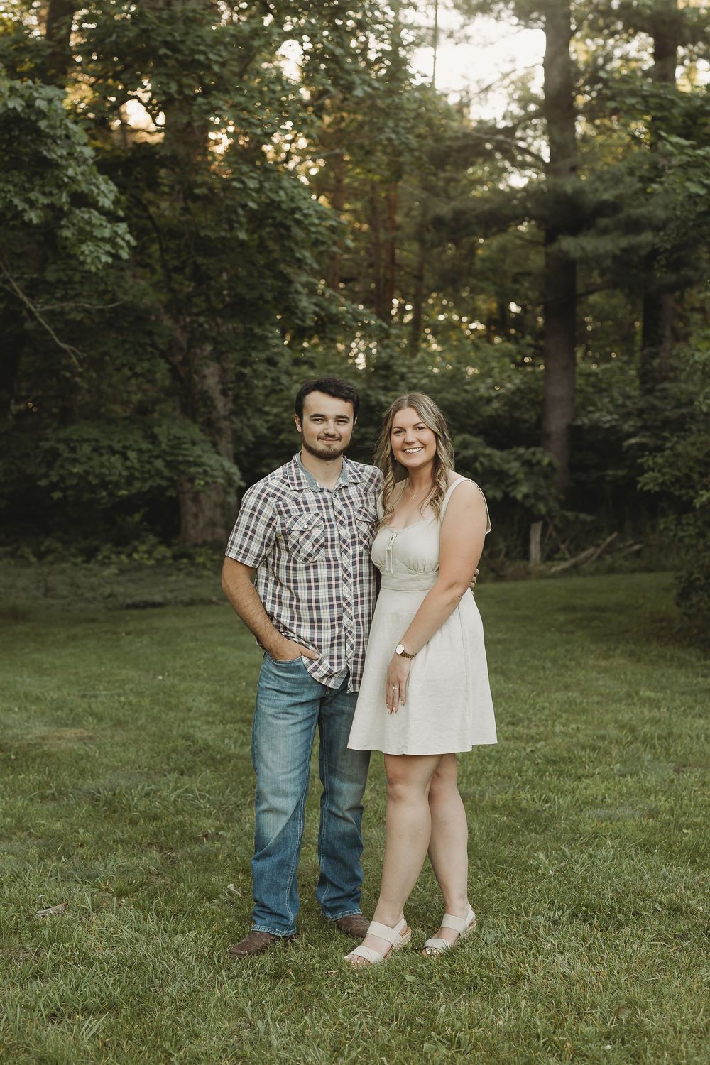 A young couple smiles together on a green lawn surrounded by lush trees at golden hour.
