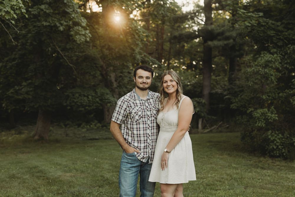 Couple smiling together in a lush green park at golden hour, man in plaid shirt and woman in white dress.