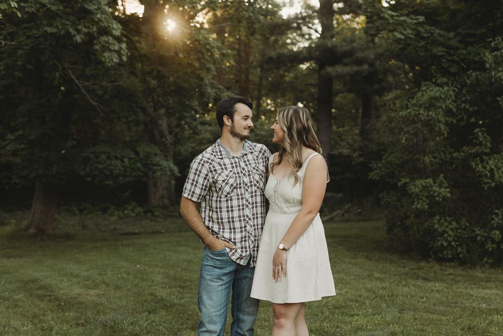 Couple smiling at each other in a lush green park during golden hour sunset, man in plaid shirt and woman in white dress.