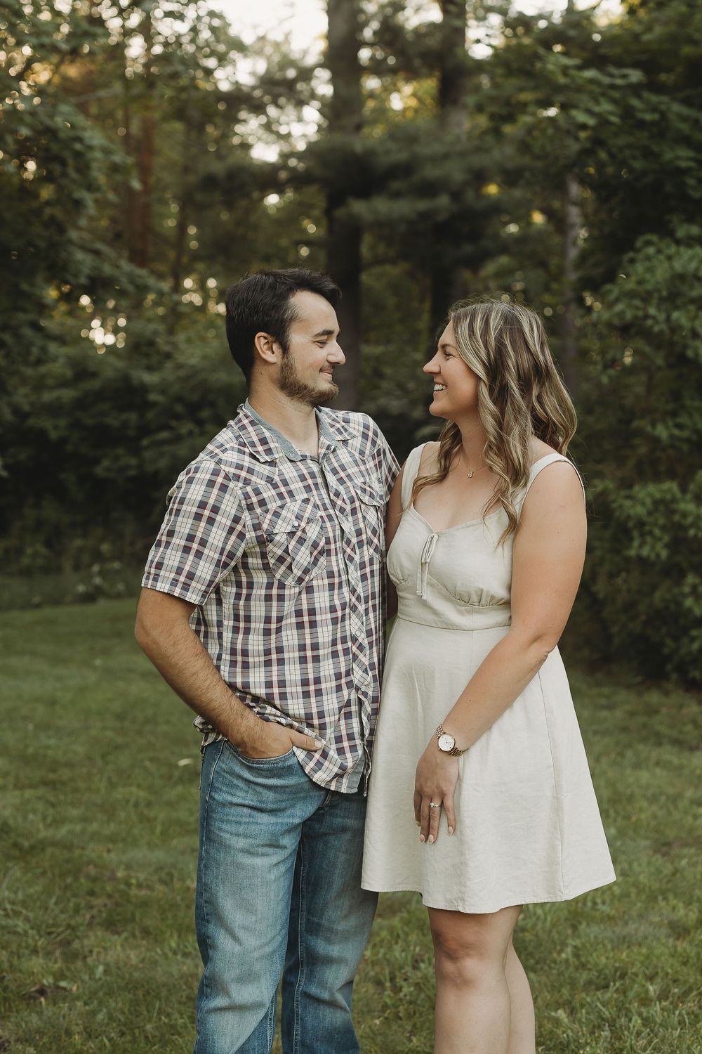 A couple smiles at each other outdoors, man in plaid shirt and jeans, woman in white dress, green trees behind.