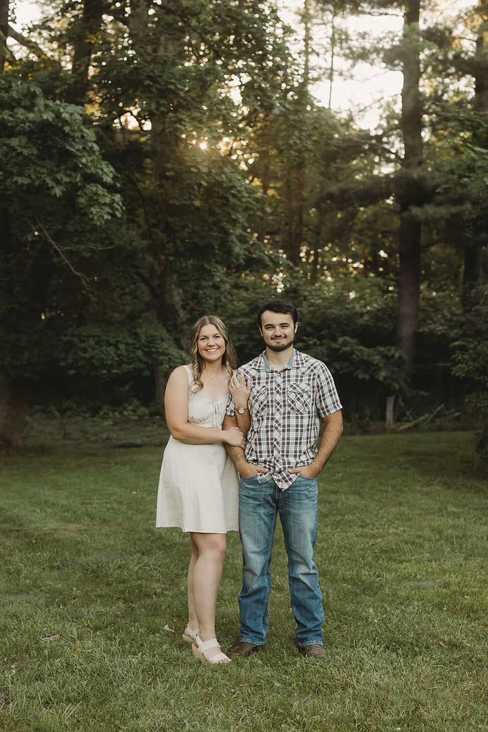 A smiling couple poses together on a green lawn surrounded by trees during golden hour.