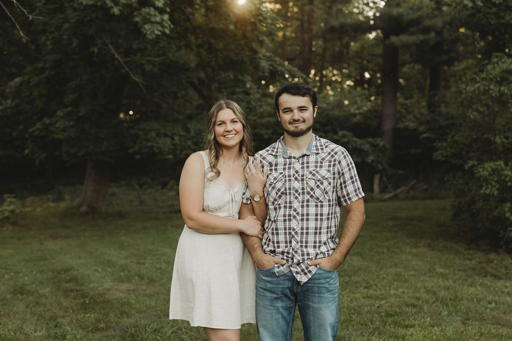 Couple smiling together outdoors at golden hour, woman in white dress and man in plaid shirt, lush green trees background.