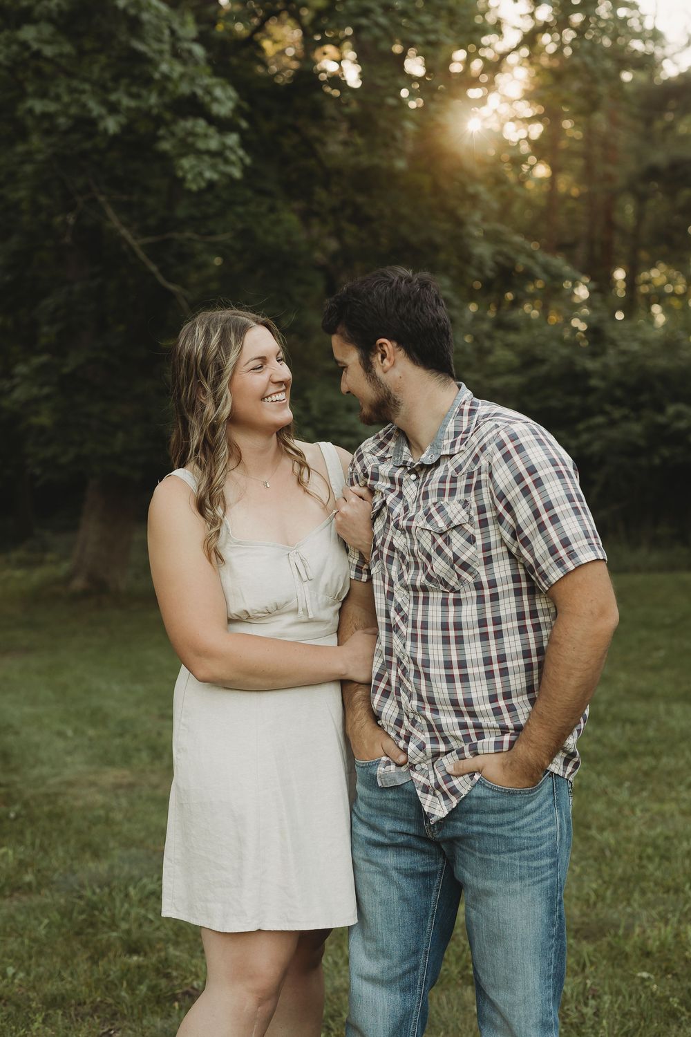 A smiling couple shares a tender moment outdoors at golden hour, woman in white dress, man in plaid shirt and jeans.