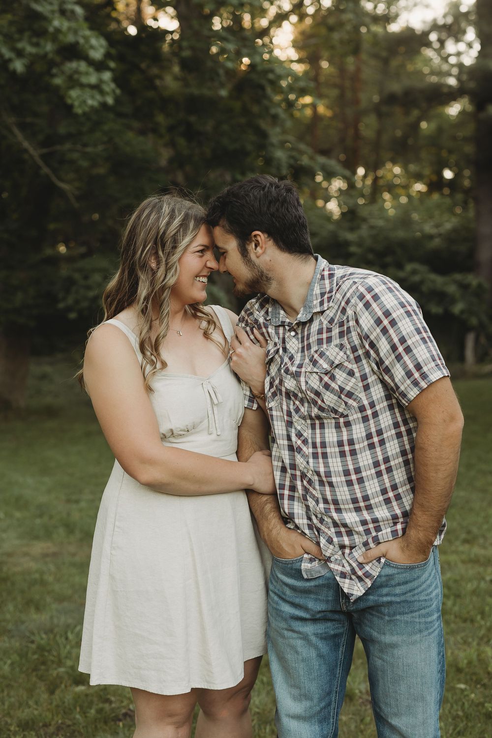 Couple sharing a tender moment outdoors, woman in white dress and man in plaid shirt, smiling together in a green park.