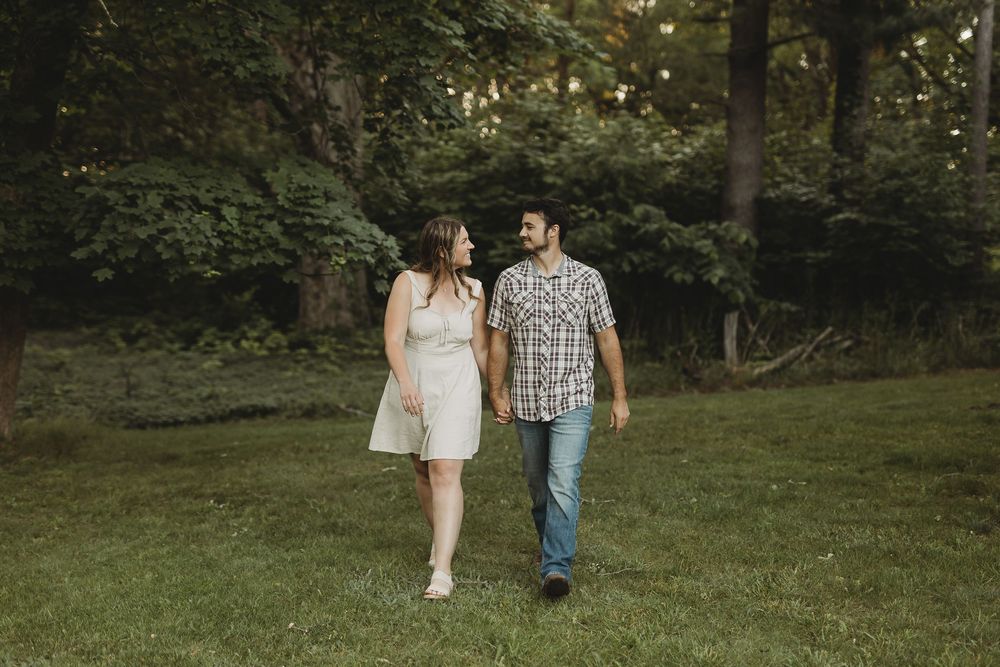 Couple holding hands walking through lush green garden, woman in white dress and man in plaid shirt and jeans.