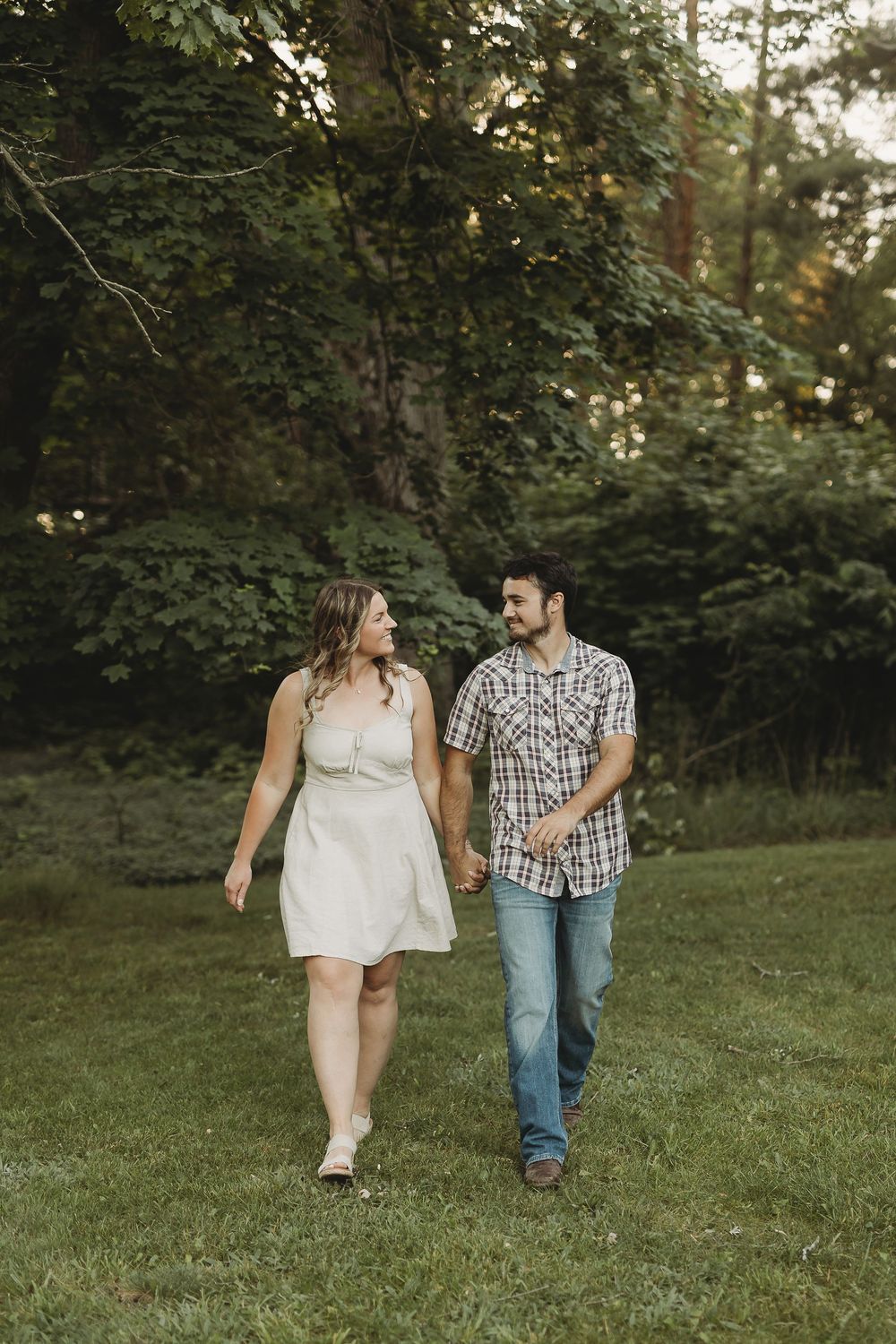A couple holds hands and walks through a lush green park, smiling at each other, woman in white dress, man in plaid shirt.