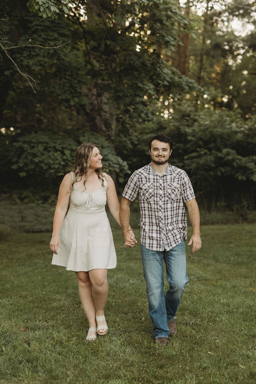 A couple walks hand-in-hand through a lush green park, woman in white dress, man in plaid shirt and jeans.