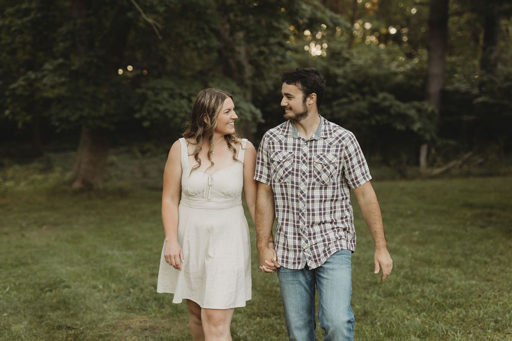 A smiling couple walks hand-in-hand through a lush green park, the woman in a white dress and man in a plaid shirt.
