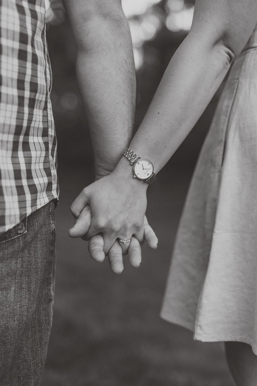 Black and white photo of a couple holding hands, woman wearing a watch and ring, man in plaid shirt and jeans.