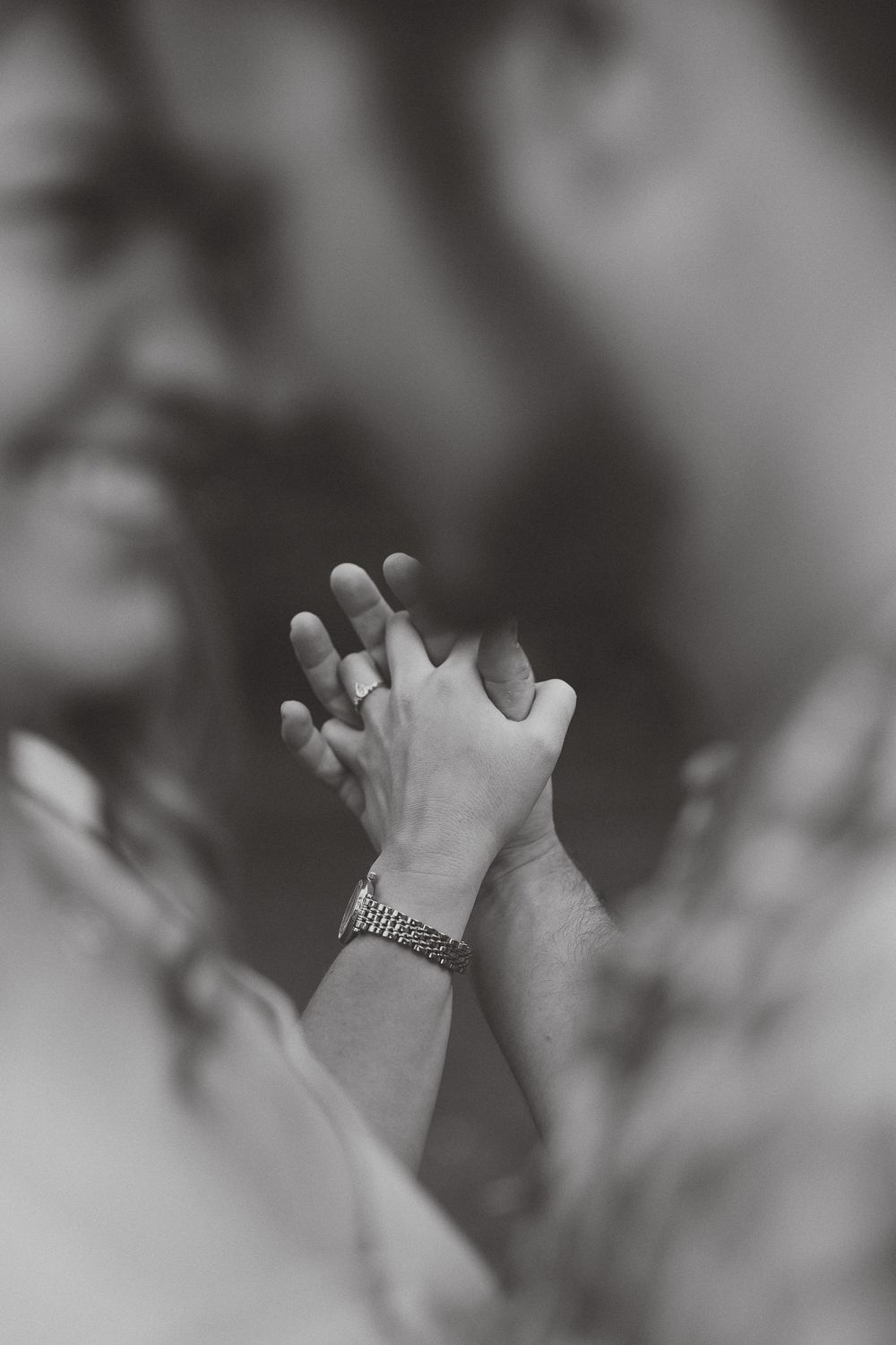 Black and white photo of couple holding hands, woman wearing ring, man with bracelet, blurred faces in background.