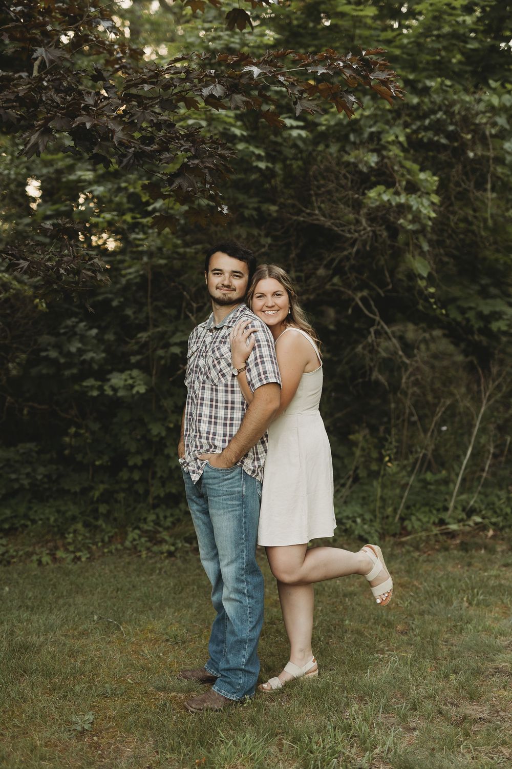 A smiling couple poses outdoors in a garden, woman in white dress hugging man in plaid shirt and jeans.
