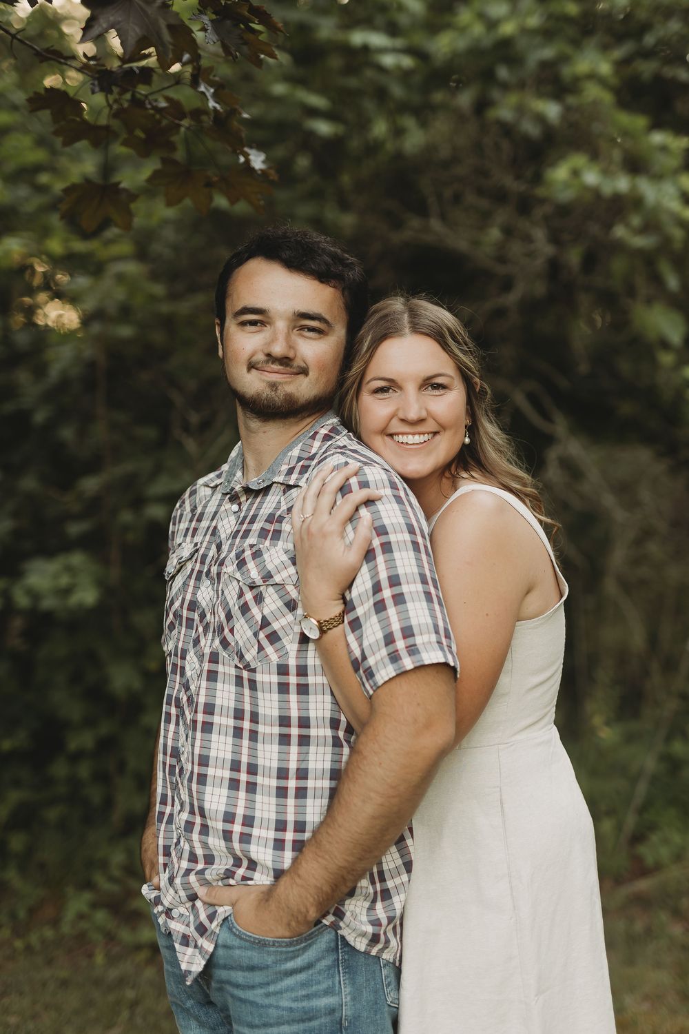 A smiling couple poses outdoors near green trees, woman in white dress embracing man in plaid shirt.
