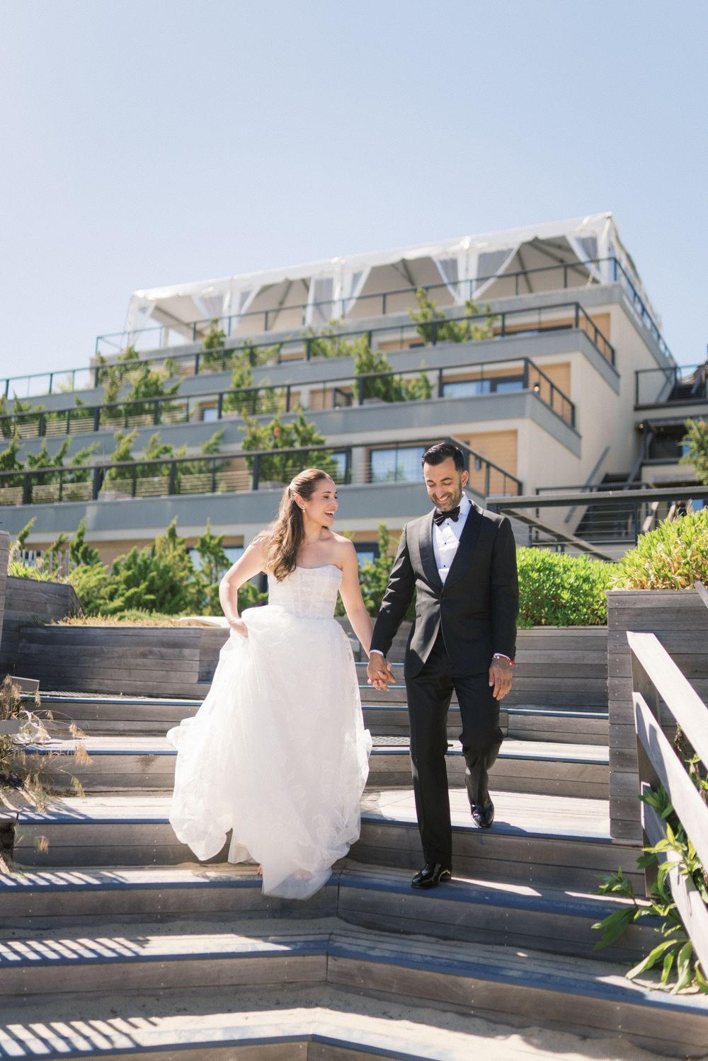 Bride and groom walk hand-in-hand down outdoor steps at a modern venue on a sunny day.