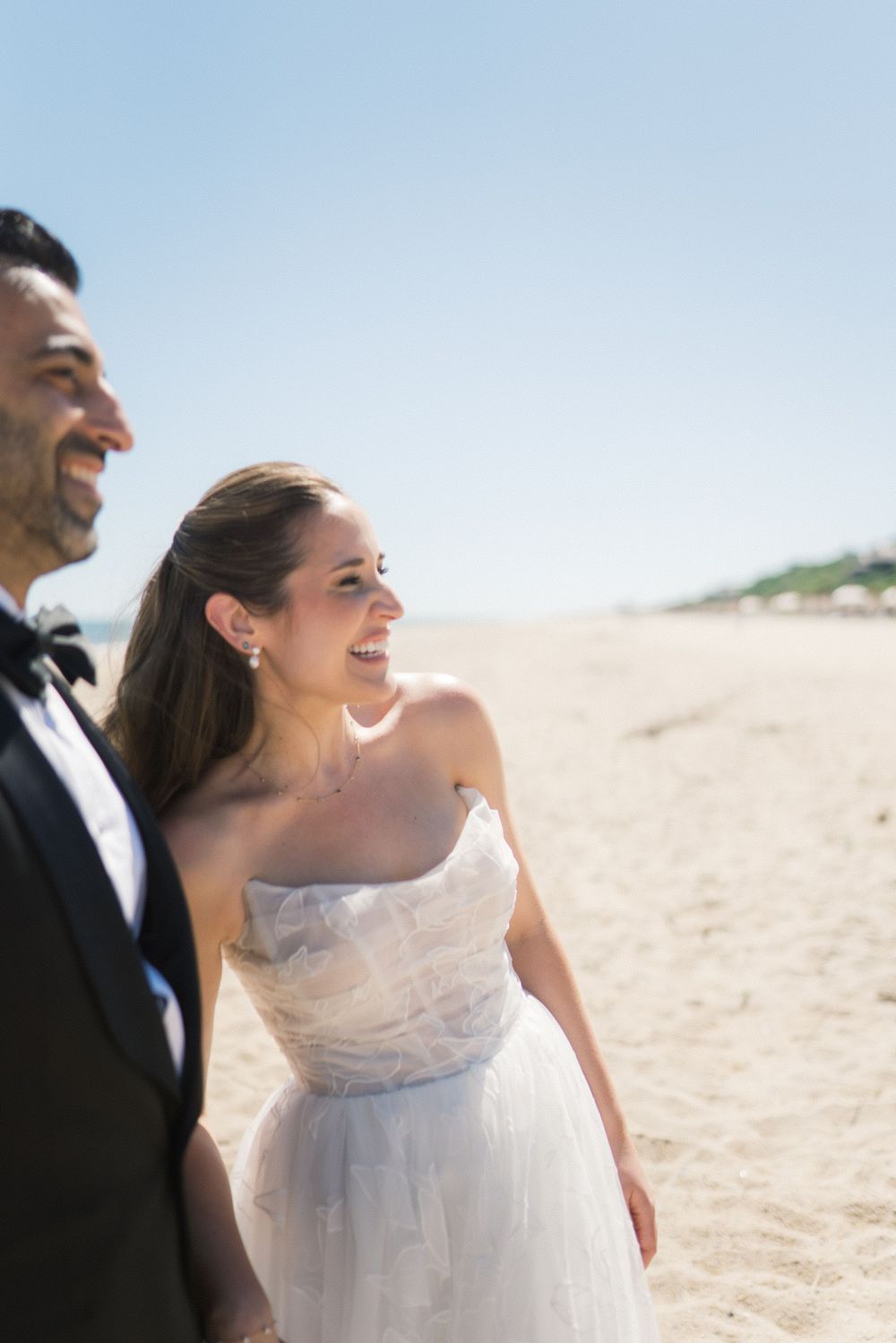 Smiling bride in strapless white gown and groom in tuxedo walking on sandy beach on sunny wedding day.