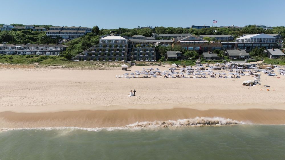 Aerial view of a sandy beach with lounge chairs, calm ocean waves, and a hotel resort surrounded by lush green trees.