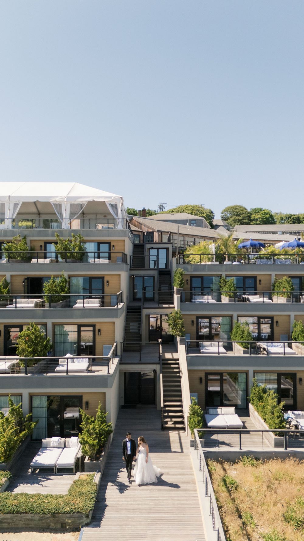 Aerial view of a wedding couple walking on a boardwalk at a modern multi-level hotel with balconies and lush greenery.