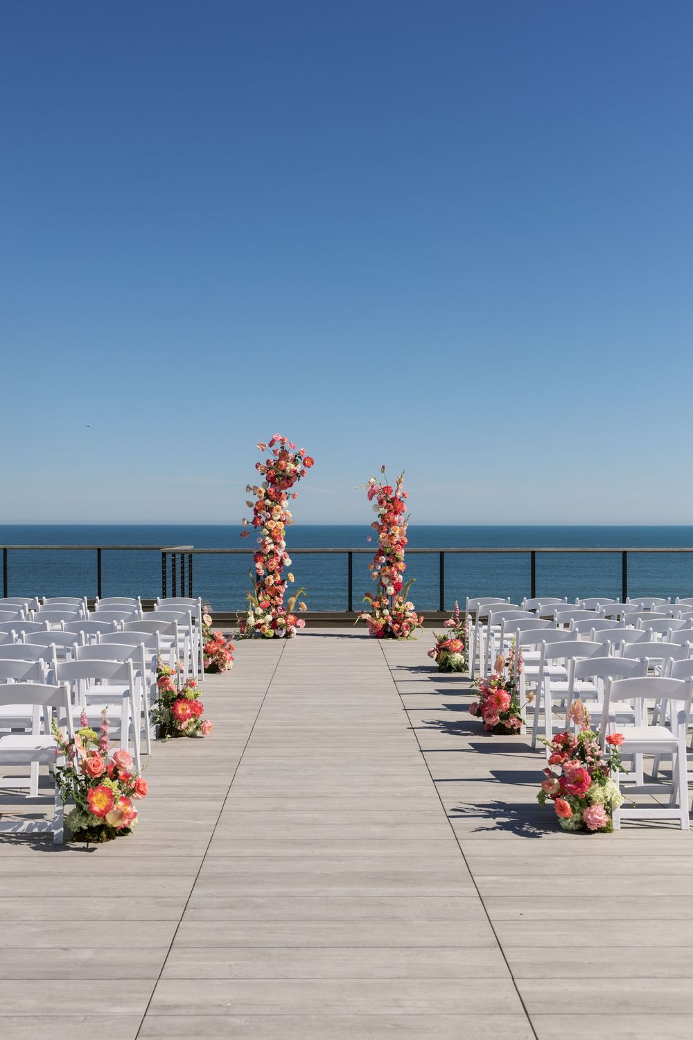 Outdoor oceanfront wedding ceremony setup with white chairs, colorful floral columns, and a clear blue sky backdrop.