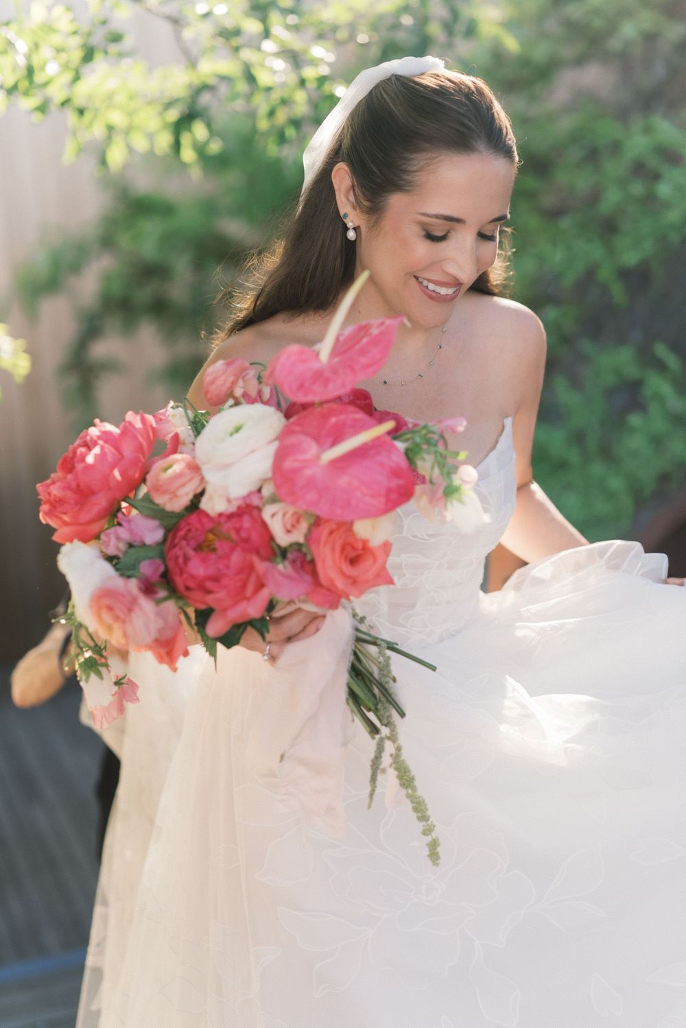 Smiling bride in white lace gown holding vibrant pink and coral bouquet with peonies and anthuriums on her wedding day.