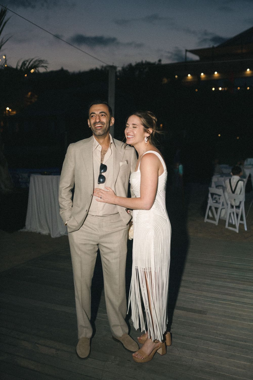 Couple in elegant beige and white outfits smiling at an outdoor evening wedding reception on a wooden deck.