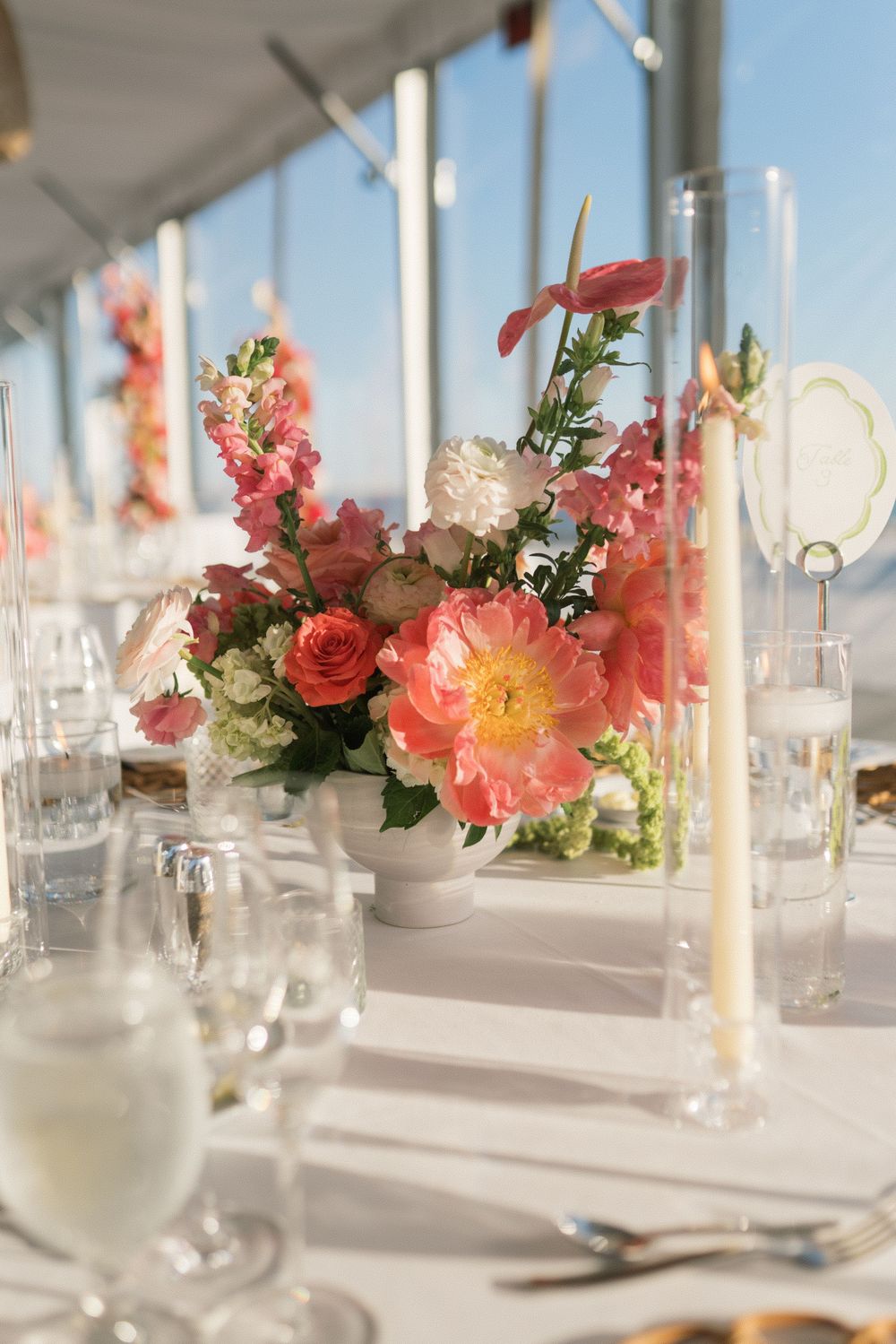 Elegant wedding reception table with pink peony floral centerpiece, candles, and glassware under a white tent.