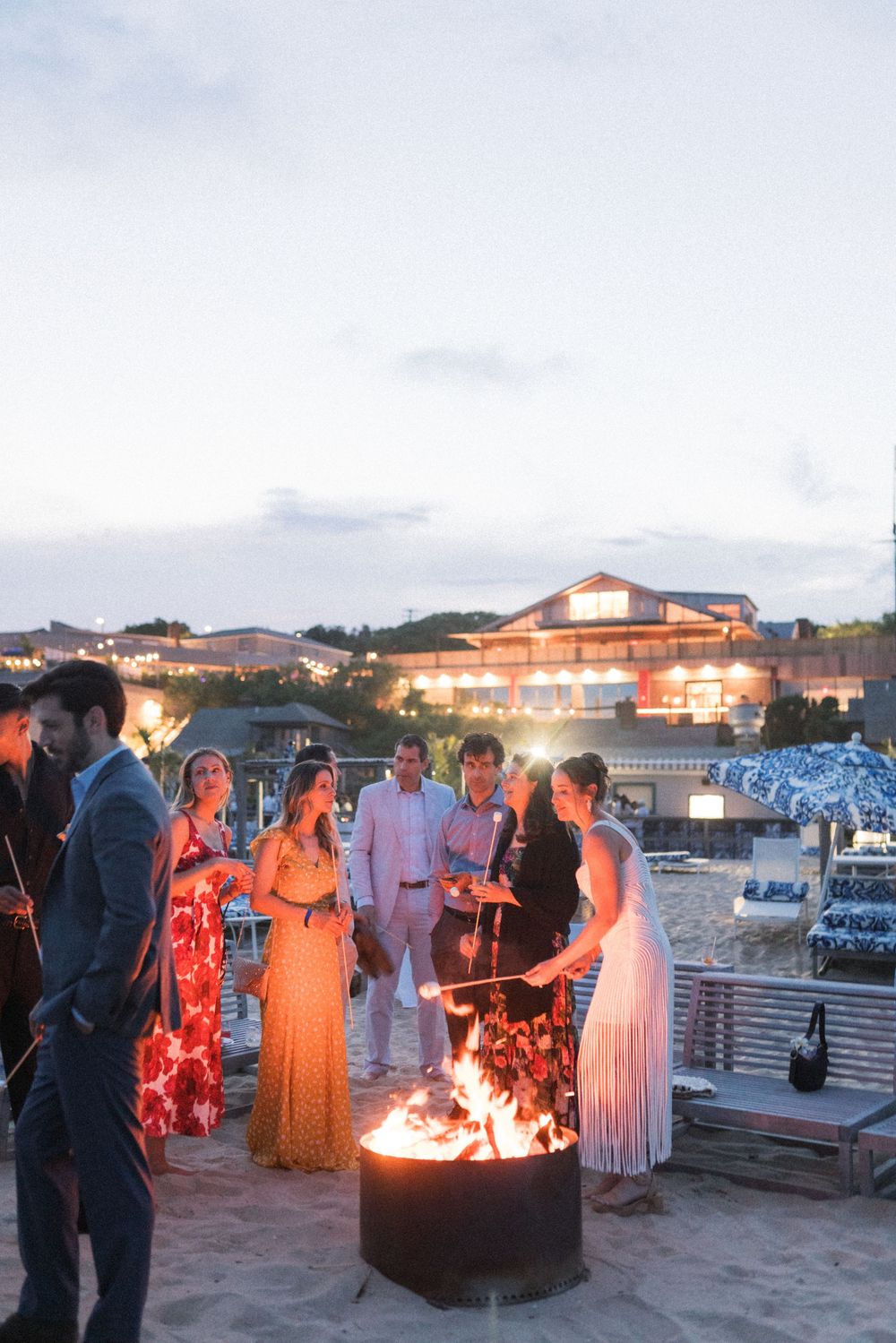 Guests gather around a beach bonfire at dusk during an outdoor wedding reception with a lit venue in the background.