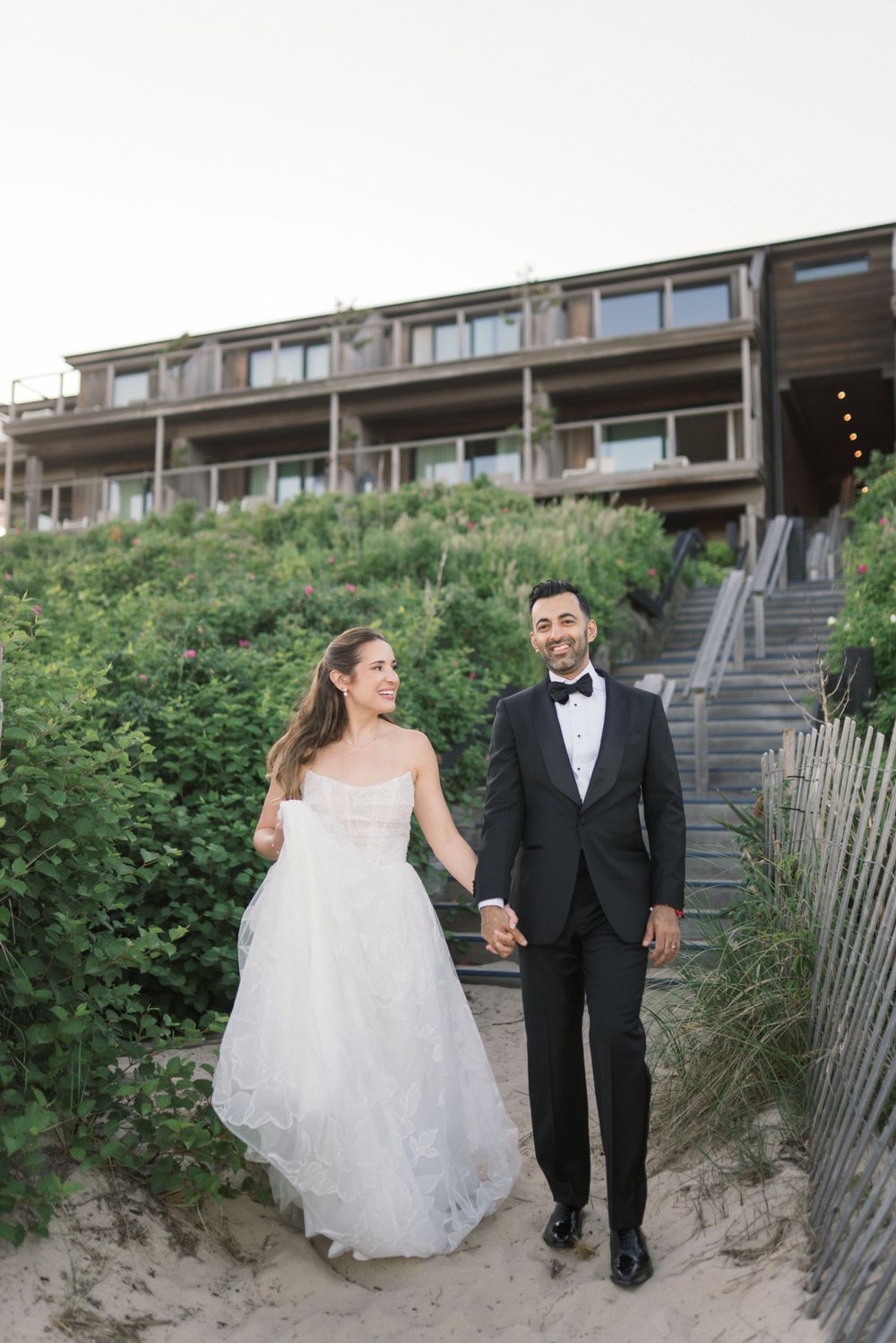 Bride and groom holding hands on sandy dunes in front of a beachside resort at dusk.