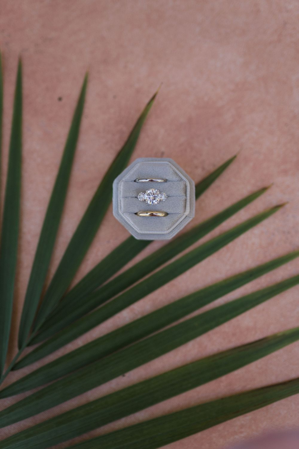 Gold wedding ring set with diamond engagement ring displayed in gray velvet box on tropical palm leaf.