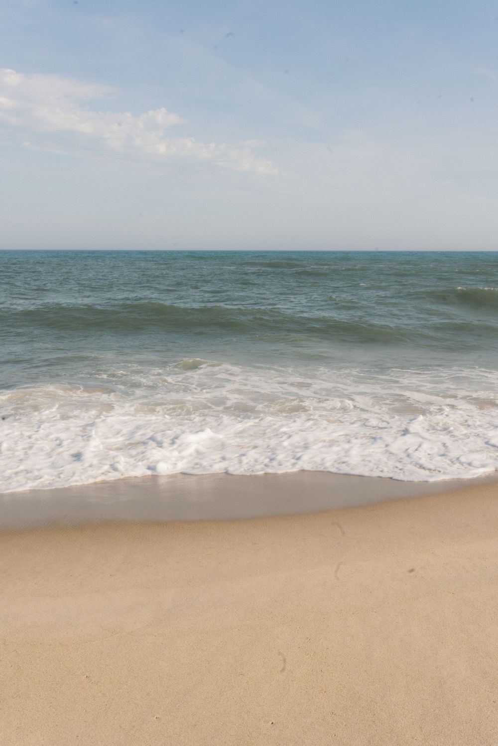 Gentle ocean waves washing onto a sandy beach under a partly cloudy blue sky.